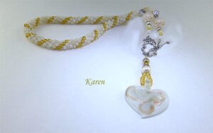 collier de perles blanches avec insertion de perles or, il est terminé par un embout de métal en argent, une grosse perle et deux petites perles son fermoir est décoratif et a bascule. il a un pendentif en forme de coeur blanc avec des insertions de couleur or. Sa taille est de 60 cm incluant le fermoir Le pendentif en verre mesure 4 1/2 cm long