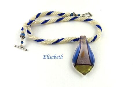 collier de perles Elisabeth fait avec des perles blanches et insertion de perles bleue le collier est terminé par un petit embout de métal ensuite deux perles blanches et une bleue. Ce collier est terminé avec un fermoir a bascule qui est décoratif. Son pendentif est de couleur argent, bleu et vert.