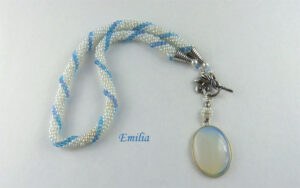 Collier de perles Emilia Collier de perles Émilia.Un collier qui sort du lot. Des petites perles blanches avec insertion de perles bleues .la terminaison du collier est avec des cônes argent et de petites perles bleues et blanc Fermoir est a bascule et aussi très décoratif. Idéal pour les personnes qui ont des douleurs aux mains. Sa longueur est de 47 cm Le pendentif est en forme ovale avec les couleurs de bleues et blanche il mesure 4 cmwww.metiersdart-cadeaux.com