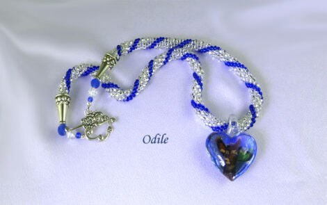 Collier de perles Odlie. Collier fait avec des perles blanches et insertions de perles de verre bleu. Ce collier est terminé par un cône de métal argent, et des petites perles blanches et bleues Il mesure 55 centimètres. Son pendentif en forme de coeur est bleu avec une tête d'animal il fait 4 centimètres.