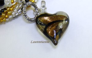 Collier de perles Laurentienne, Sa conception se compose de perles de verre transparentes avec insertion de perles or et noires le tout terminé par en embout de métal et une perle noire, transparente et jaune. Son fermoir argent très décoratif et a bascule il mesure 48 cm. Magnifique pendentif en forme de coeur de couleur or noir et transparent il mesure 3 cm 1/2 pendentif est de 3 cm 1/2