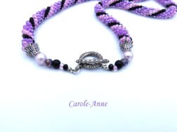 Collier crocheté avec perles de verre de couleurs mauve, rose et noires. La pièce de verre central est une création unique d’une artiste québécoise, il a la forme de A soit deux bandes de lilas et la barre du A est noire. Le collier est terminé  Le fermoir a bascule mesure 55 cm Longueur 53 cm ce collier est crocheté et terminé avec un embout de métal argent et de petites perles blanches, et noires, un fermoir a bascule rond argent termine le tout.