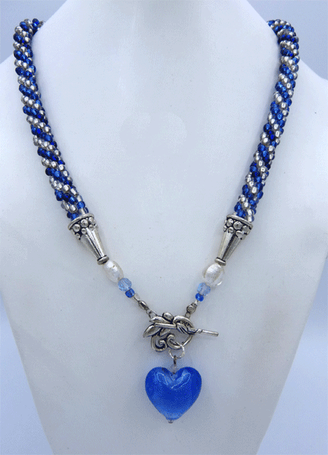 Le collier fait environ 60 cm de long. Il est fait de perles de rocaille bleue et verre transparent. Son pendentif mesure 3 cm