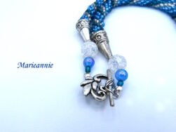 Technique de crochet de perle. Collier fait de perles de verre bleu avec insertion de perles bleu pâle et bleu foncé et une rangée de perles de verre transparente.Le bout du collier est fini par des embouts de métal et une grosse perle transparente et une autre plus petite bleue. Le fermoir est a bascule et argent il représente les pétales d'une fleur.Le pendentif  est une création québécoise avec un rectangle de bleu différent.