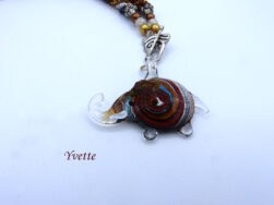 Collier de perles de verre. Il est composé de perles brunes avec insertion de perles or, blanches et bleu et un embout en métal argent .Le fermoir est en argent à bascule. Longueur52 cm incluant le fermoir Le pendentif;est une pièce de verre en forme d'éléphant, il mesure  4 cm x 5cm.