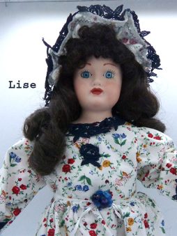 Poupée de porcelaine, ses cheveux sont bruns frisés et ses yeux bleus. Sa tête et les membres sont en porcelaine son corps est en tissus. Sa robe est blanche avec des fleurs jaunes, bleus, rouge et elle a de la dentelle bleue marine au collet de sa robe, aux manches et en bas de la robe.