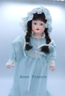 Poupée de porcelaine, ses cheveux sont bruns avec des nattes et ses yeux verts. Sa tête et les membres sont en porcelaine son corps est en tissus. Sa robe est bleue pâle , elle a de la dentelle blanche au collet de sa robe, aux manches et en bas de la robe ainsi qua la taille. Elle possède son ourson