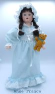 Poupée de porcelaine, ses cheveux sont bruns avec des nattes et ses yeux verts. Sa tête et les membres sont en porcelaine son corps est en tissus. Sa robe est bleue pâle , elle a de la dentelle blanche au collet de sa robe, aux manches et en bas de la robe ainsi qua la taille. Elle possède son ourson