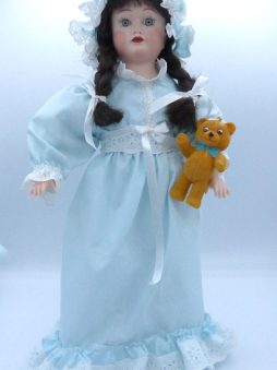 Poupée de porcelaine, ses cheveux sont bruns avec des nattes et ses yeux verts. Sa tête et les membres sont en porcelaine son corps est en tissus. Sa robe est bleue pâle , elle a de la dentelle blanche au collet de sa robe, aux manches et en bas de la robe ainsi qua la taille. Elle possède son ourson
